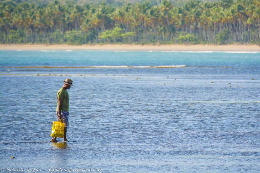 Imagem de um homem andando com uma sacola nas águas da praia.